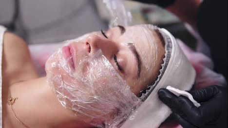 Professional-cosmetologist-is-cleaning-woman's-face-from-special-treatment-using-cotton-sponge.-Young-woman-is-lying-on-the