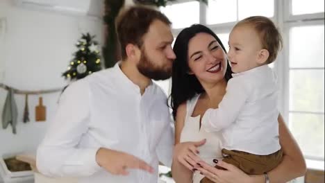 Happy-young-family-dance-together-in-their-stylish-apartment-by-the-christmas-tree-holding-their-adorable-child-in-hands.-Lovely