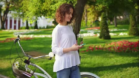 Happy-smiling-woman-messaging-using-her-smartphone-standing-in-the-city-park-near-her-city-bicycle-with-flowers-in-its-basket