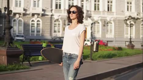 Smiling-attractive-woman-in-sunglasses-walking-in-the-old-city-street-holding-her-longboard-in-the-morning-during-the-dawn.-Slow