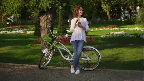 Beautiful-woman-standing-in-the-city-park-near-her-city-bicycle-with-flowers-in-its-basket-and-drinking-green-detox-smoothie