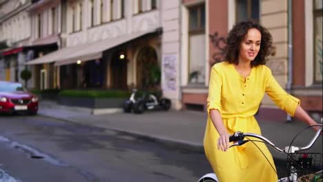 Beautiful-woman-riding-a-city-bicycle-with-a-basket-and-flowers-in-the-city-center-while-talking-to-someone.-Steadicam-shot