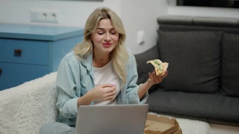 Girl-sitting-on-sofa-at-modern-living-room-and-taking-tasty-pizza-while-use-laptop