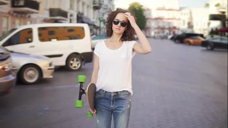 Happy-woman-touching-her-sunglasses-then-starting-to-walk-holding-her-stylish-longboard-in-the-morning.-Slow-motion-shot
