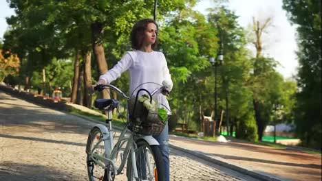 Young-woman-in-a-white-t-shirt-and-blue-jeans-walking-on-the-cobblestone-road-in-the-city-park-holding-her-city-bicycle