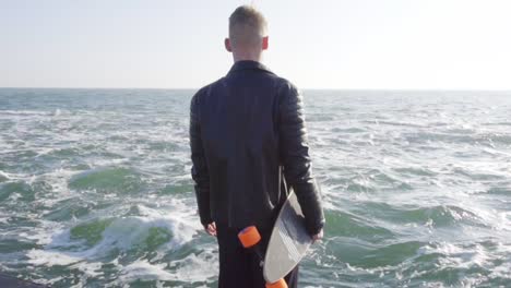 Young-man-in-black-leather-jacket-holds-his-longboard-by-the-sea