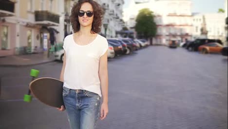 Happy-woman-in-sunglasses-walking-in-the-old-city-street-holding-her-stylish-longboard-in-the-morning.-Slow-motion-shot
