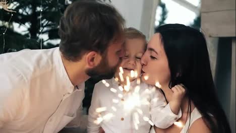 Beautiful-family-rejoices-the-new-year-and-lights-sparklers,-young-parents-kissing-their-lovely-child-sitting-by-the-christmas