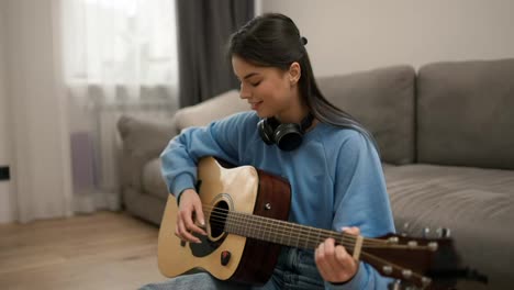 Young-girl-learning-to-play-the-guitar,-sitting-on-the-floor-in-headphones