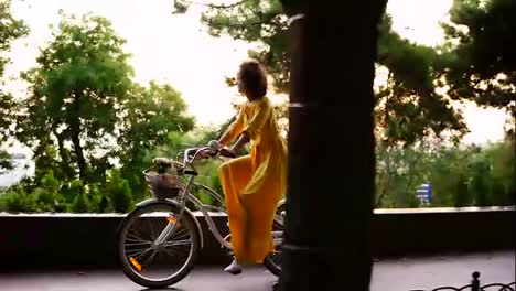 Young-brunette-woman-in-long-yellow-dress-cycling-with-flowers-and-packet-in-a-basket-and-exploring-the-city.-Steadicam-shot