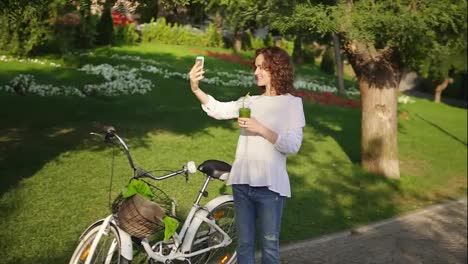 Portrait-of-a-young-woman-taking-selfie-photo-with-cell-phone-standing-in-the-city-park-near-her-city-bicycle-with-flowers-in-its