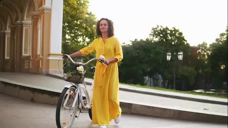 Smiling-young-girl-in-a-long-yellow-dress-walking-holding-her-city-bicycle-handlebar-with-flowers-in-its-basket-in-the-city