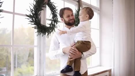 Young-father-is-holding-his-baby-boy,-standing-by-the-window-and-looking-outside.-The-window-is-decorated-with-Christmas-wreath