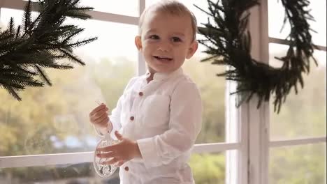 Adorable-blonde-toddler-in-white-shirt-standing-on-the-window-sill-holding-a-christmas-toy-and-smiling.-The-window-is-decorated