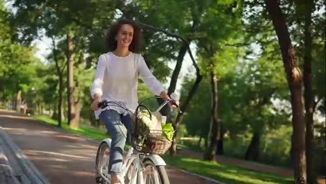 Happy-smiling-woman-is-enjoying-her-time-riding-a-city-bicycle-with-a-basket-and-flowers-inside-during-the-dawn.-Steadicam-shot