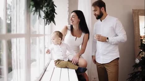 Young-lovely-family-sitting-together-by-the-window-decorated-with-Christmas-wreath.-Loving-parents-kissing-each-other-in-the