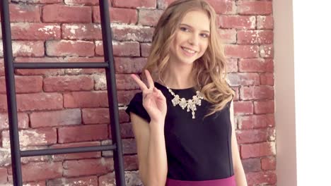 Bright-young-blond-girl-is-smiling-and-posing-against-the-brick-wall-showing-v-sign-victory-or-peace
