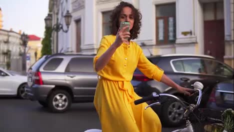 Beautiful-smiling-woman-in-long-yellow-dress-riding-a-city-bicycle-with-a-basket-and-flowers-inside-and-filming-using-her-phone