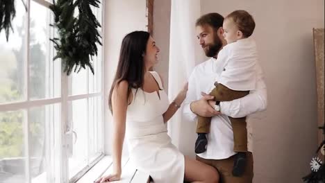 Young-father-is-holding-his-baby-boy,-standing-by-the-window-while-attractive-mother-in-white-dress-is-sitting-on-the-window