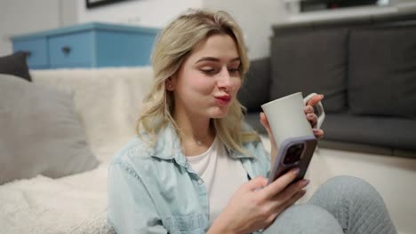 Smiling-girl-is-messaging-online-on-the-phone,-holds-mug-with-hot-drink,-checks-emails