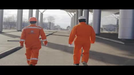 Dos-Trabajadores-De-La-Construcción-Felices-Con-Uniforme-Naranja-Y-Cascos-Caminando-Y-Saltando-Juntos