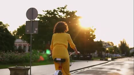 Vista-Posterior-De-Una-Mujer-Irreconocible-Con-Un-Largo-Vestido-Amarillo-Montando-Una-Bicicleta-En-El-Centro-De-La-Ciudad-Durante-El-Amanecer-Disfrutando-De-Ella