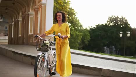Attractive-happy-woman-in-a-long-yellow-dress-walking-in-the-city-holding-her-city-bicycle-handlebar-with-flowers-in-its