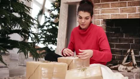 Beautiful-smiling-woman-wrapping-Christmas-presents-sitting-by-the-Christmas-tree-and-fireplace-at-home