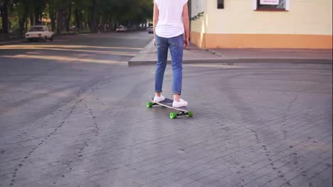 Back-view-of-a-woman-in-a-white-t-shirt,-blue-jeans-and-white-sneakers-skateboarding-in-the-city-street.-Camera-moves-from-legs