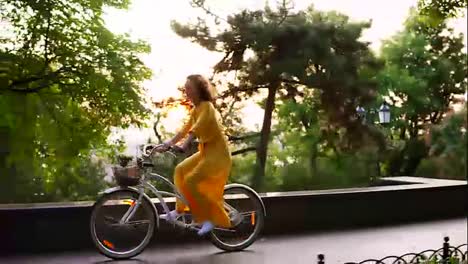 Beautiful-smiling-woman-in-long-yellow-dress-riding-a-city-bicycle-with-a-basket-and-flowers-inside-during-the-dawn.-Lens-flare