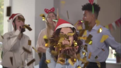 Young-attractive-woman-blowing-golden-confetti-from-hands-wearing-christmas-hat-while-her-co-workers-partying-in-the-background