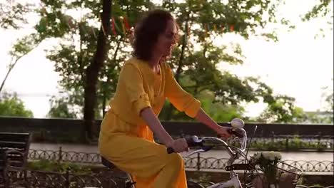 Attractive-woman-in-long-yellow-dress-is-cycling-through-the-city-in-summertime-on-the-bike-with-basket-and-flowers-in-the