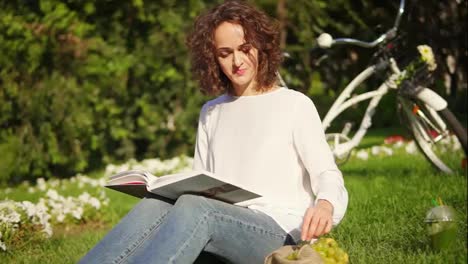 Young-woman-in-white-shirt-and-blue-jeans-is-reading-a-book-sitting-on-the-grass-in-park-and-taking-grapes.-Her-city-bicycle