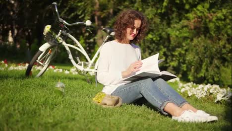 Attractive-woman-in-white-shirt-and-blue-jeans-is-reading-a-book-sitting-on-the-grass-in-park-during-sunny-warm-day.-Her-city