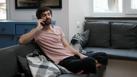 Smiling-bearded-man-calling-a-person,-listening-and-talking-with-smartphone-in-home-living-room