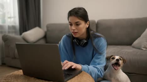 Young-woman-in-headphones-sitting-on-floor-with-her-dog-working-on-laptop