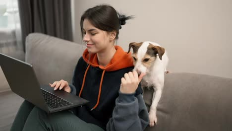 Young-woman-sitting-on-a-couch-working-on-laptop-with-her-dog-next-her