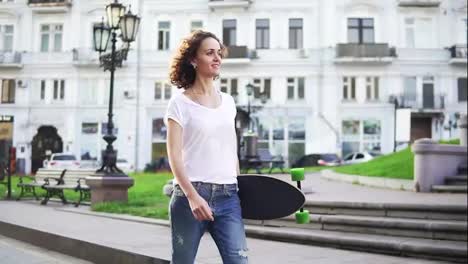 Smiling-attractive-girl-walking-in-the-old-city-street-holding-her-longboard-in-the-morning,-slow-motion-shot.-Beautiful-old