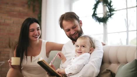 Young-father-and-his-son-reading-book,-while-mother-is-drinking-tea-and-kissing-her-husband.-Family-reading-Christmas-story