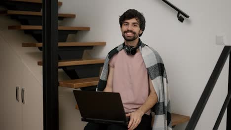 Smiling-bearded-man-with-laptop,-sitting-on-wooden-stairs-at-home-in-plaid