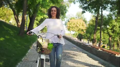 Smiling-attractive-woman-in-a-white-t-shirt-and-blue-jeans-walking-holding-her-city-bicycle-handlebar-with-flowers-in-its