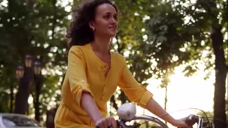 Lens-flare:-smiling-happy-woman-in-long-yellow-dress-is-riding-a-city-bicycle-with-a-basket-and-flowers-in-the-park-with-green