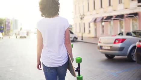 Back-view-of-a-woman-walking-in-the-old-city-street-holding-her-trendy-stylish-longboard-with-flowers-in-the-morning.-Lens-flare
