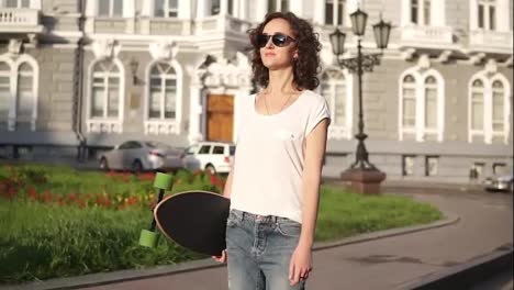 Beautiful-young-woman-in-sunglasses-walking-in-the-old-city-street-holding-her-longboard-in-the-morning-during-the-dawn