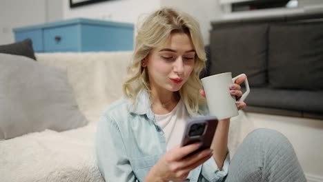 Blonde-girl-is-messaging-online-on-the-phone,-holds-mug-with-hot-drink,-checks-emails