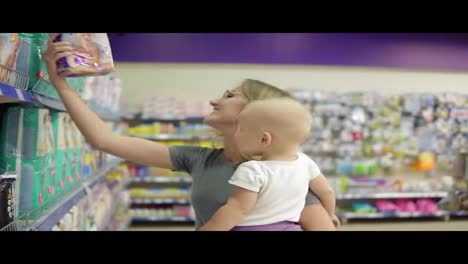 Smiling-mother-in-a-section-for-children-in-the-supermarket-holding-her-child-in-her-arms-while-choosing-diapers-on-the-shelves