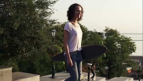 Young-attractive-girl-walking-holding-her-longboard-in-the-city-street-during-sunrise,-slow-motion-shot.-Beautiful-city-harbor