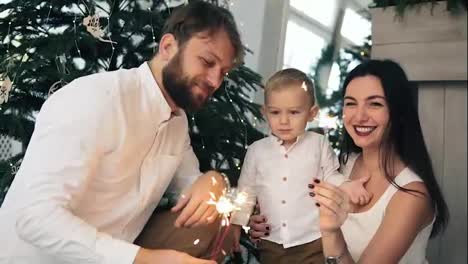 Mother-and-father-holding-sparkler,-while-their-cute-child-is-looking-at-light.-Happy-family-and-Bengal-fire-at-home-by-the