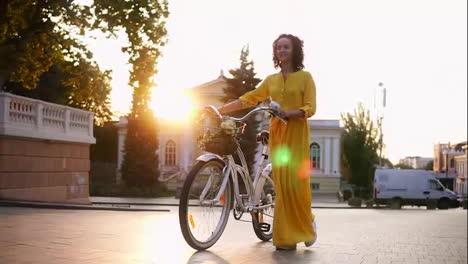 Beautiful-young-woman-in-a-long-yellow-dress-walking-during-the-dawn-holding-her-city-bicycle-handlebar-with-flowers-in-its