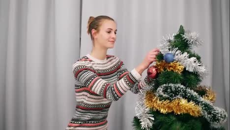 Preparation-for-Christmas:-attractive-happy-woman-sitting-by-the-artificial-christmas-tree-and-decorating-it-at-home